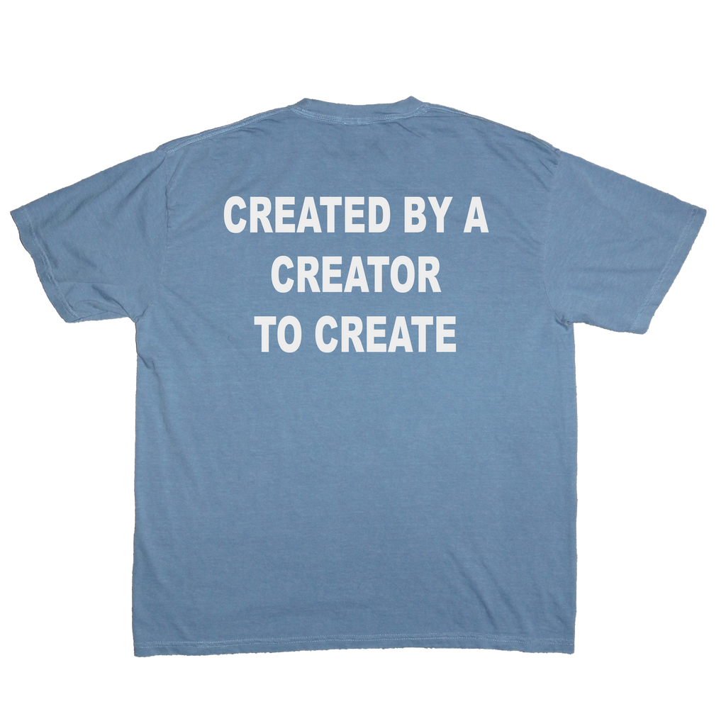 A light blue crewneck t-shirt shown from the back, featuring the phrase 'CREATED BY A CREATOR TO CREATE' printed in bold white capital letters across the upper back.