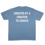 A light blue crewneck t-shirt shown from the back, featuring the phrase 'CREATED BY A CREATOR TO CREATE' printed in bold white capital letters across the upper back.