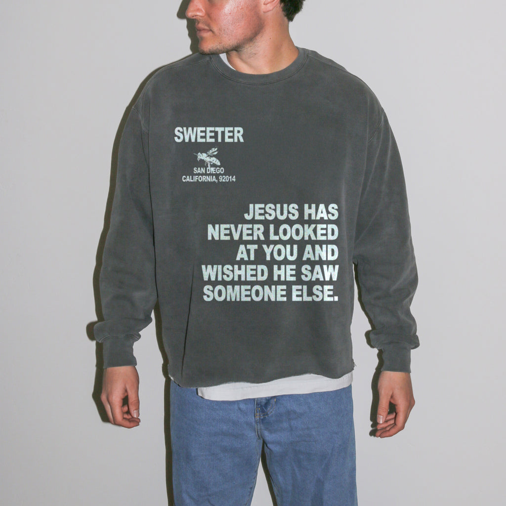 A man wearing a dark gray washed sweatshirt with white text that reads 'SWEETER SAN DIEGO CALIFORNIA 9214' and 'JESUS HAS NEVER LOOKED AT YOU AND WISHED HE SAW SOMEONE ELSE.', paired with blue jeans against a plain light gray background.