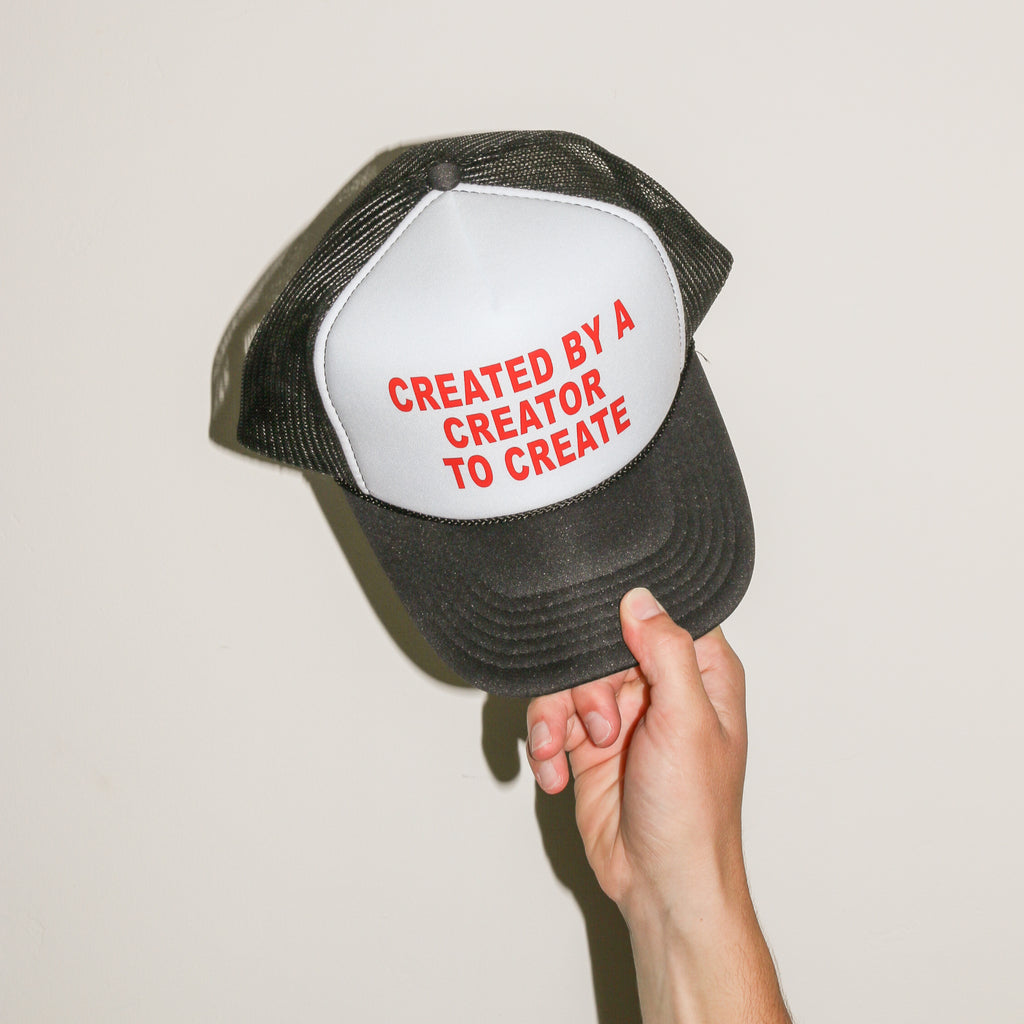 A hand holding a black and white trucker hat with red embroidered text that reads 'CREATED BY A CREATOR TO CREATE' against a plain off-white background.