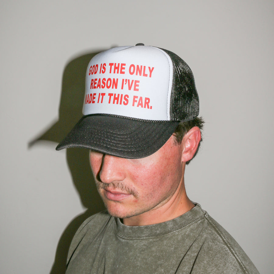 A man wearing a trucker hat with a white front panel, black mesh back, and red text that reads 'GOD IS THE ONLY REASON I'VE MADE IT THIS FAR.'.