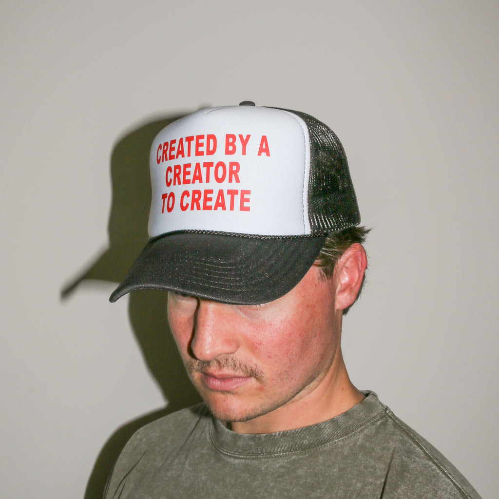 A man wearing a trucker hat with a white front panel and black mesh back, featuring the red text 'CREATED BY A CREATOR TO CREATE' on the front. The man is looking down and wearing a gray t-shirt against a plain light gray wall.