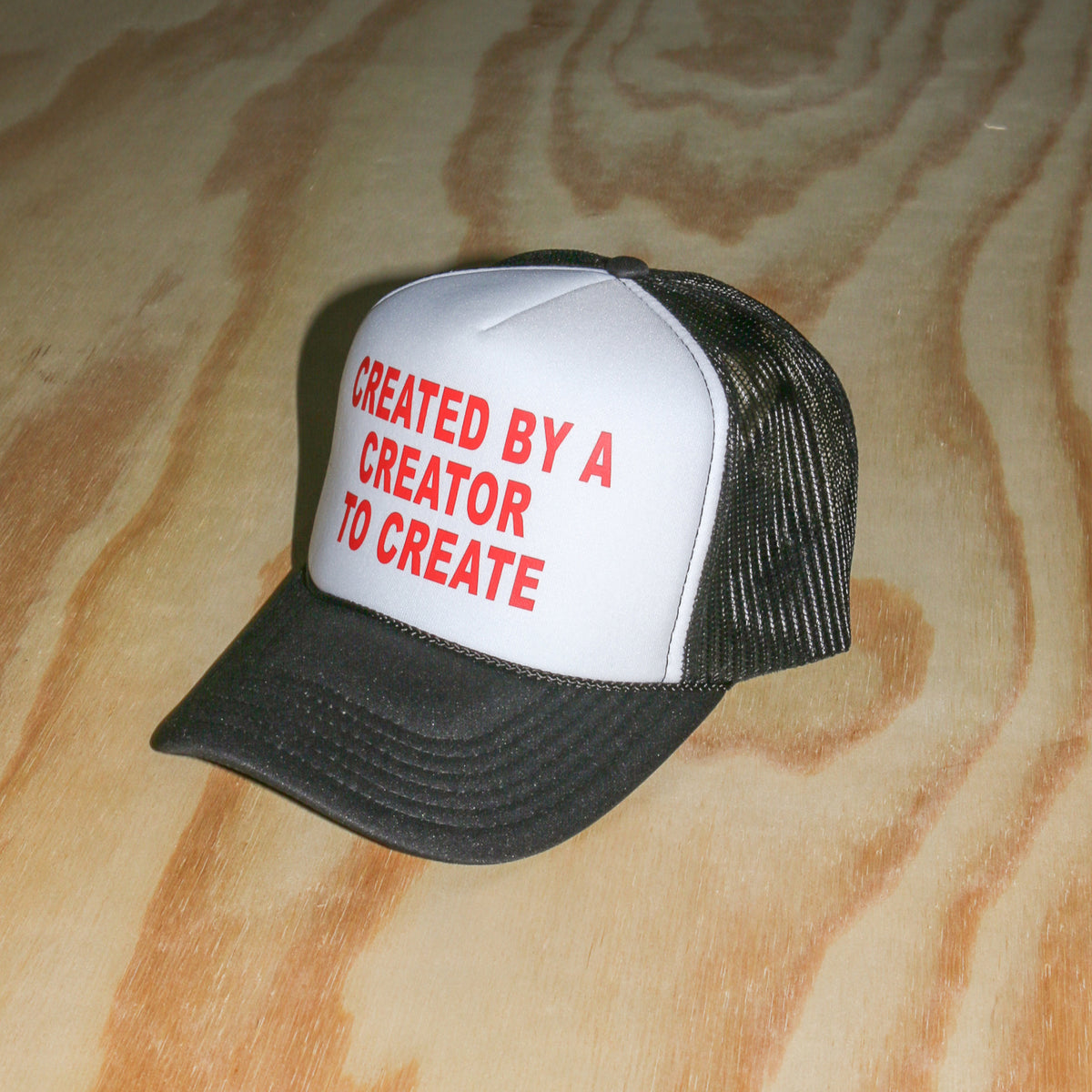 A black and white trucker hat with red text reading 'CREATED BY A CREATOR TO CREATE' resting on a light wood grain surface.