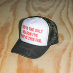 A black and white trucker hat with red text reading 'GOD IS THE ONLY REASON I'VE MADE IT THIS FAR.' resting on a light wood grain surface.