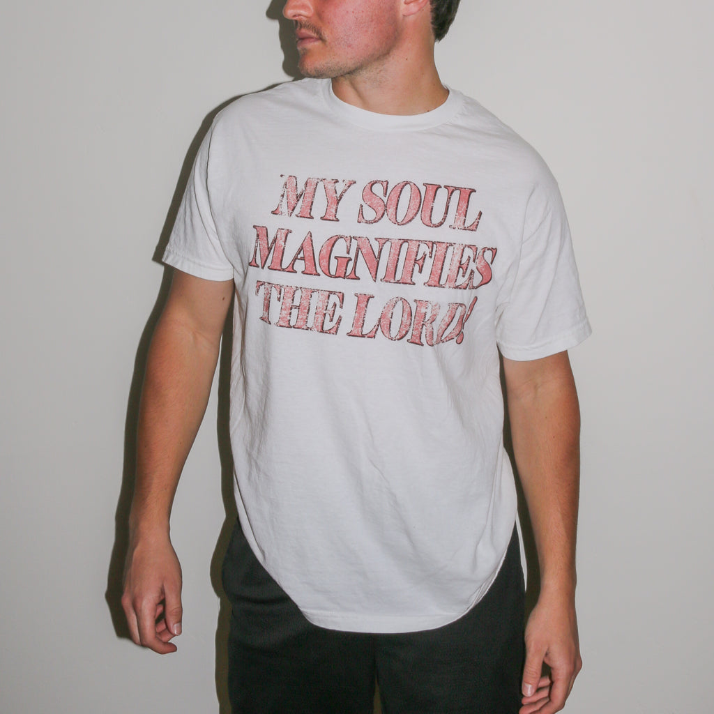 A man wearing a white crewneck t-shirt with the text 'MY SOUL MAGNIFIES THE LORD!' printed in a distressed red font, set against a plain light gray wall.