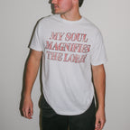 A man wearing a white crewneck t-shirt with the text 'MY SOUL MAGNIFIES THE LORD!' printed in a distressed red font, set against a plain light gray wall.