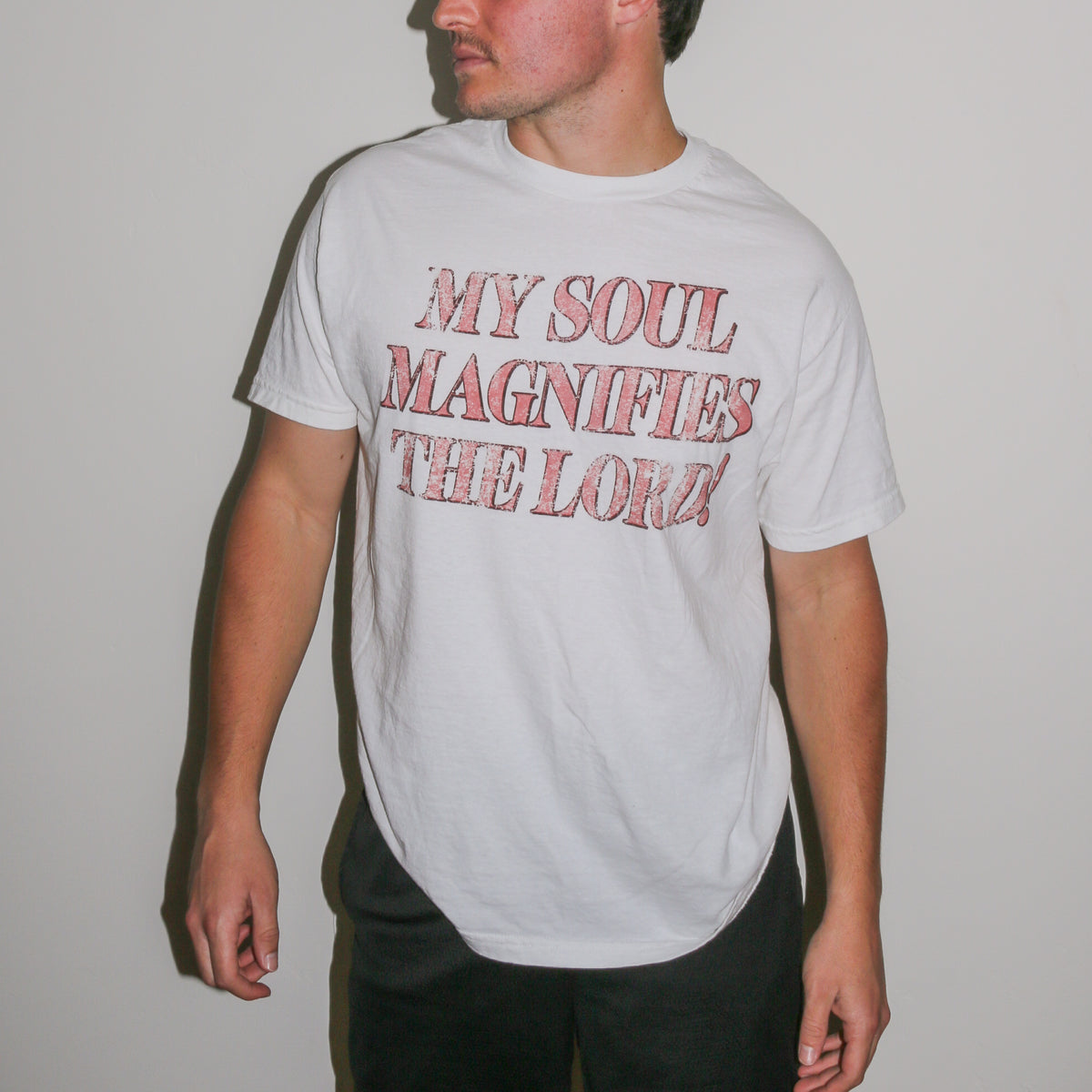 A man wearing a white crewneck t-shirt with the text 'MY SOUL MAGNIFIES THE LORD!' printed in a distressed red font, set against a plain light gray wall.