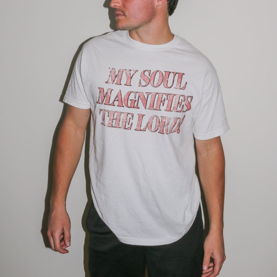 A man wearing a white crewneck t-shirt with the text 'MY SOUL MAGNIFIES THE LORD!' printed in a distressed red font, set against a plain light gray wall.
