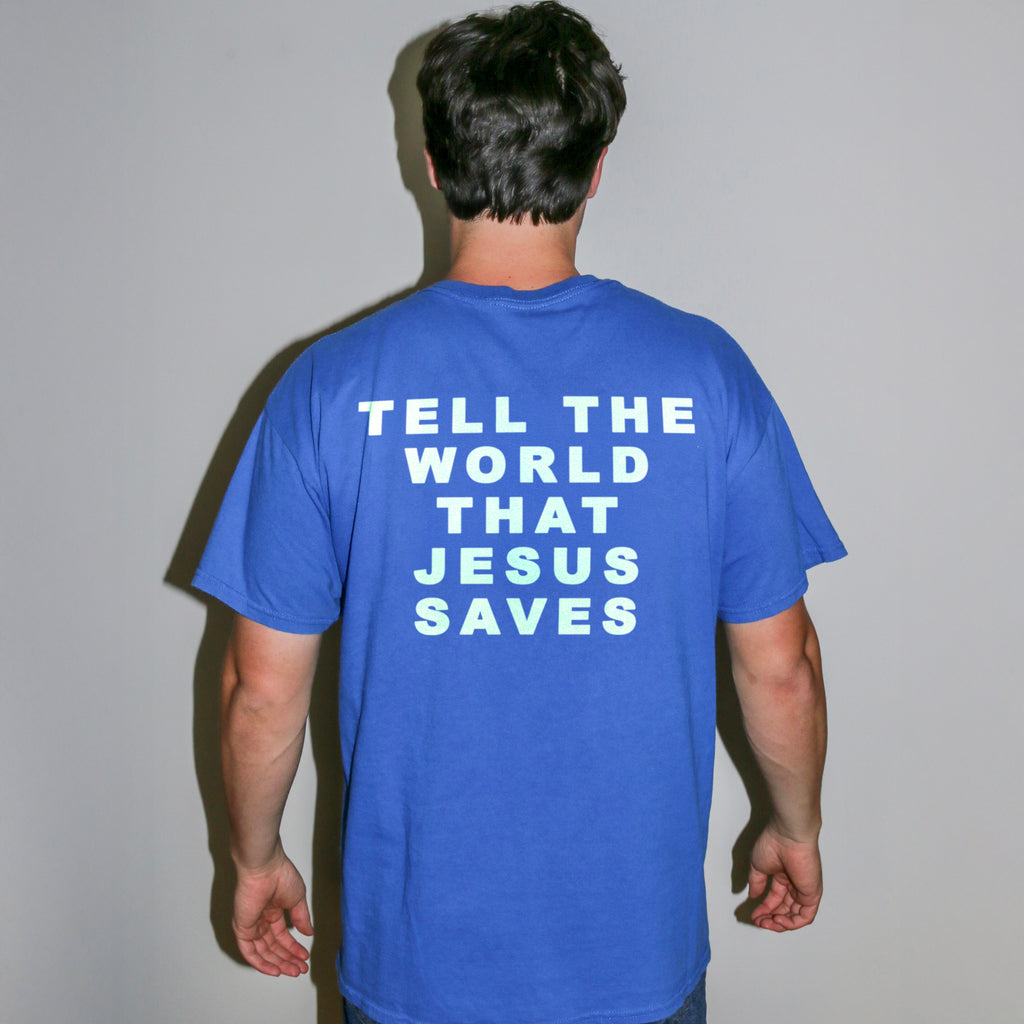 A man viewed from behind wearing a royal blue t-shirt with the slogan 'TELL THE WORLD THAT JESUS SAVES' printed in white block letters on the back, set against a plain light gray wall.