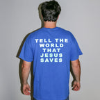 A man viewed from behind wearing a royal blue t-shirt with the slogan 'TELL THE WORLD THAT JESUS SAVES' printed in white block letters on the back, set against a plain light gray wall.