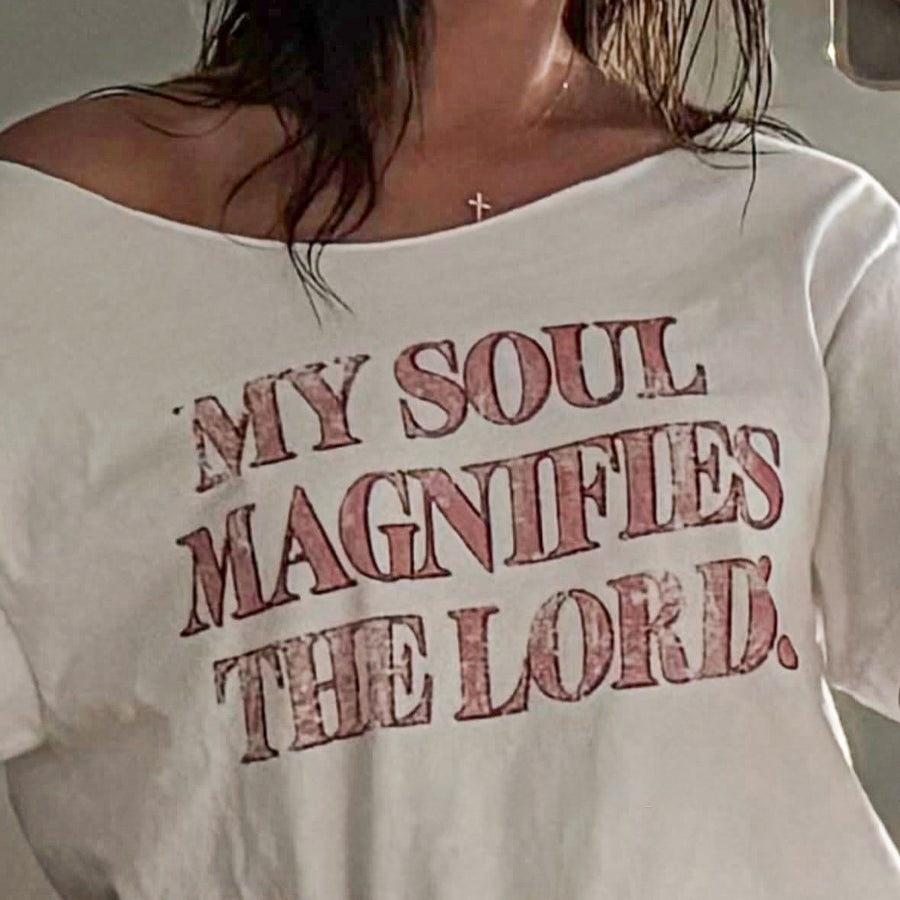 A close-up of a person wearing a white off-the-shoulder t-shirt with the text 'MY SOUL MAGNIFIES THE LORD.' printed in a distressed reddish-brown font across the chest.