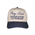 A baseball cap with a cream-colored crown and a navy blue brim, featuring embroidered text on the front that reads 'Pray Loud' and 'REJOICE ALWAYS, PRAY WITHOUT CEASING, GIVE THANKS IN ALL CIRCUMSTANCES'.