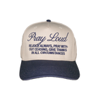 A baseball cap with a cream-colored crown and a navy blue brim, featuring embroidered text on the front that reads 'Pray Loud' and 'REJOICE ALWAYS, PRAY WITHOUT CEASING, GIVE THANKS IN ALL CIRCUMSTANCES'.