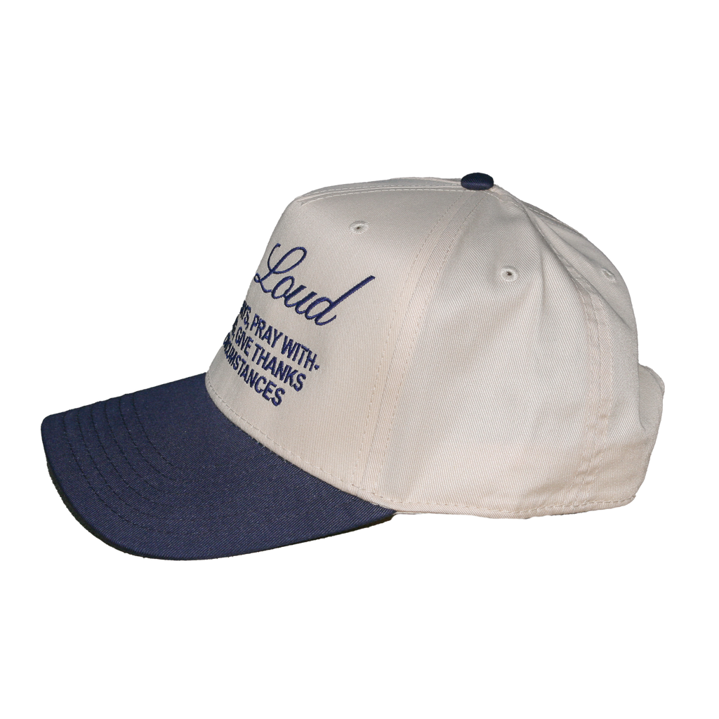A baseball cap with an off-white crown and a navy blue brim, featuring navy blue embroidered text on the front that reads 'Loud' and 'pray with thanksgiving'.