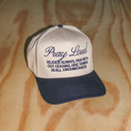 A baseball cap with a cream-colored front panel and a dark navy blue brim, featuring the embroidered text 'Pray Loud' and a scripture on the front. The cap is photographed on a light-colored wood grain surface.