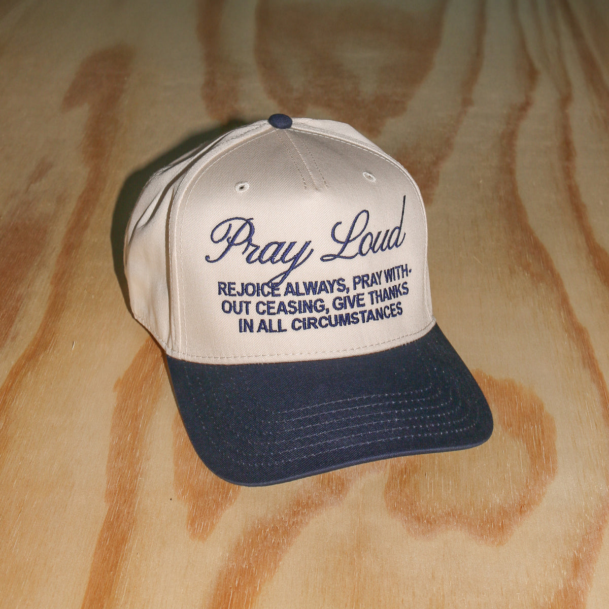 A baseball cap with a cream-colored front panel and a dark navy blue brim, featuring the embroidered text 'Pray Loud' and a scripture on the front. The cap is photographed on a light-colored wood grain surface.