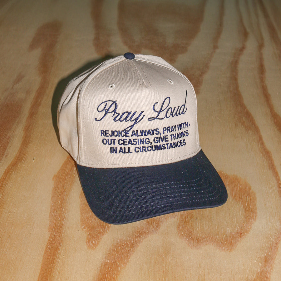 A baseball cap with a cream-colored front panel and a dark navy blue brim, featuring the embroidered text 'Pray Loud' and a scripture on the front. The cap is photographed on a light-colored wood grain surface.