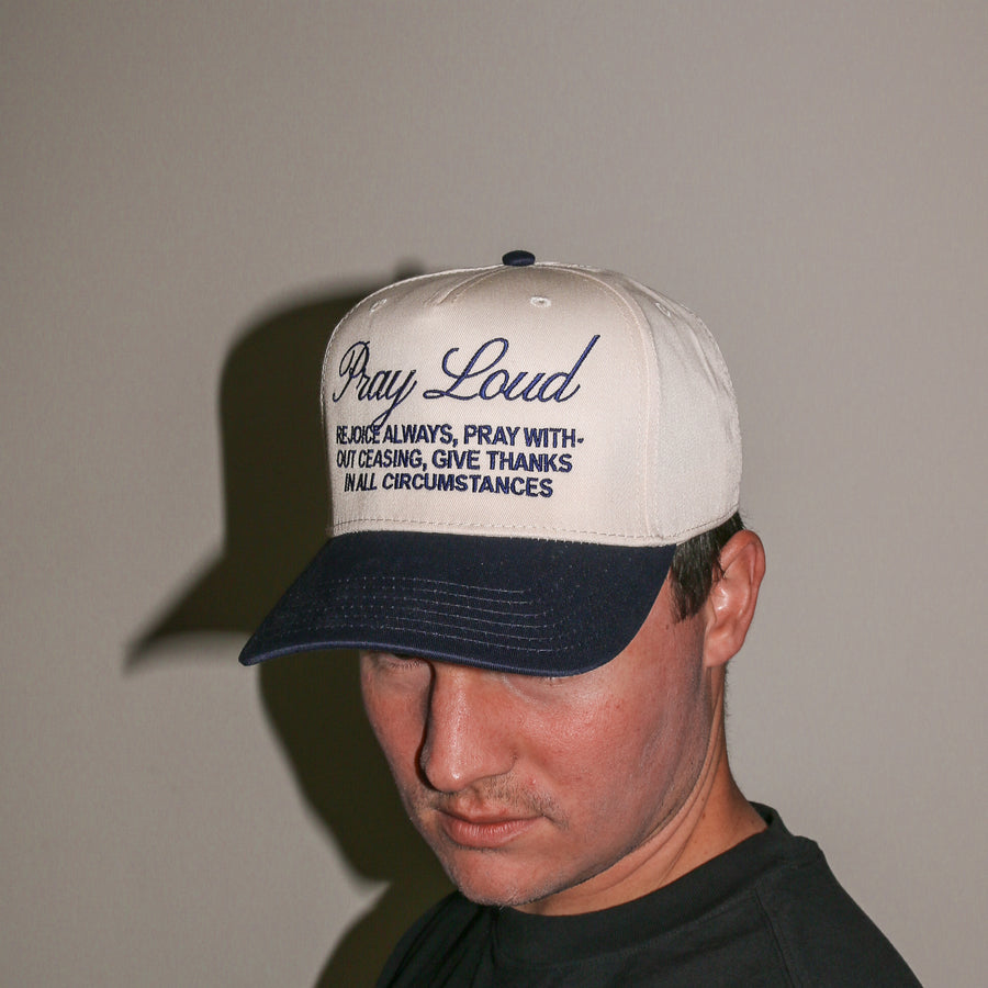 A man wearing a cream and navy blue baseball cap with embroidered text that reads 'Pray Loud' and a bible verse, photographed against a plain beige wall.