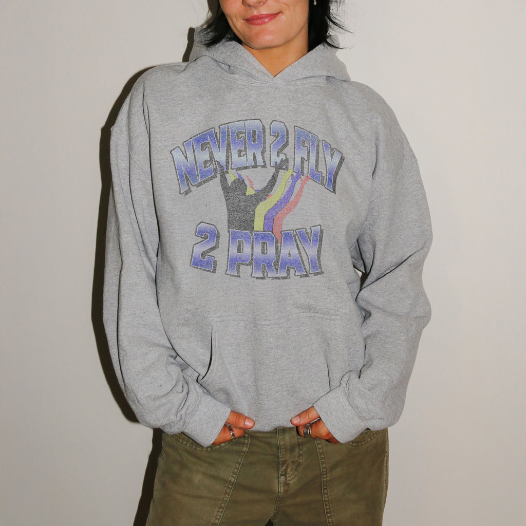 A person wearing a heather gray pullover hoodie with a graphic that reads 'NEVER 2 FLY 2 PRAY' in blue-purple lettering with a rainbow and silhouette design, paired with olive green pants against a plain light background.