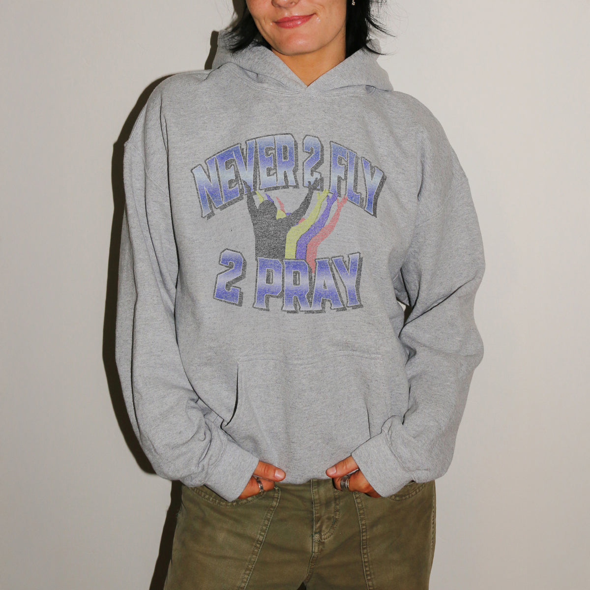 A person wearing a heather gray pullover hoodie with a graphic that reads 'NEVER 2 FLY 2 PRAY' in blue-purple lettering with a rainbow and silhouette design, paired with olive green pants against a plain light background.