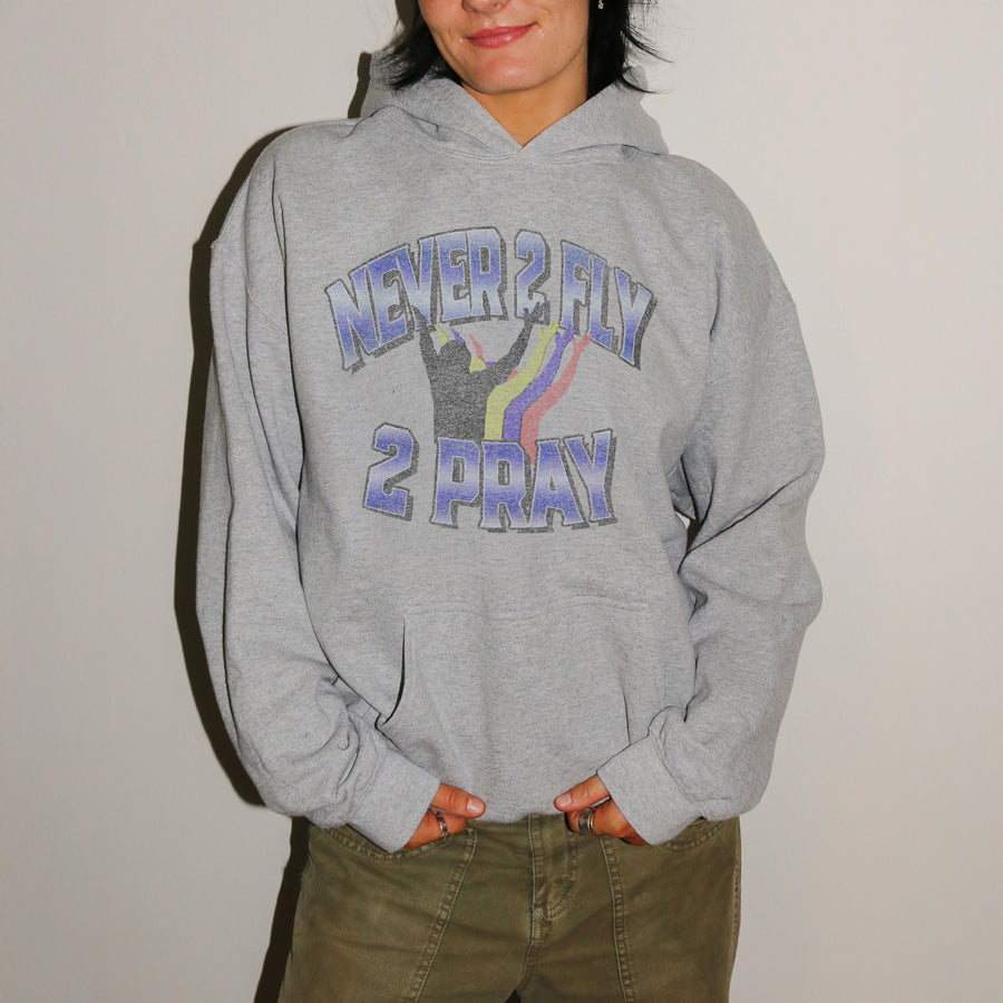 A person wearing a heather gray pullover hoodie with a graphic that reads 'NEVER 2 FLY 2 PRAY' in blue-purple lettering with a rainbow and silhouette design, paired with olive green pants against a plain light background.