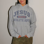 A person wearing a light gray pullover hoodie with 'JESUS UNLIMITED ATHLETIC DEPT' printed in navy and red on the front, paired with olive green pants, standing against a plain light wall.