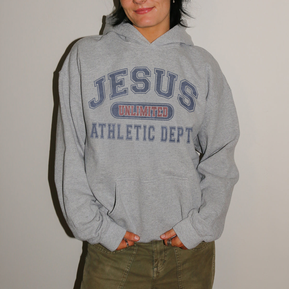 A person wearing a light gray pullover hoodie with 'JESUS UNLIMITED ATHLETIC DEPT' printed in navy and red on the front, paired with olive green pants, standing against a plain light wall.