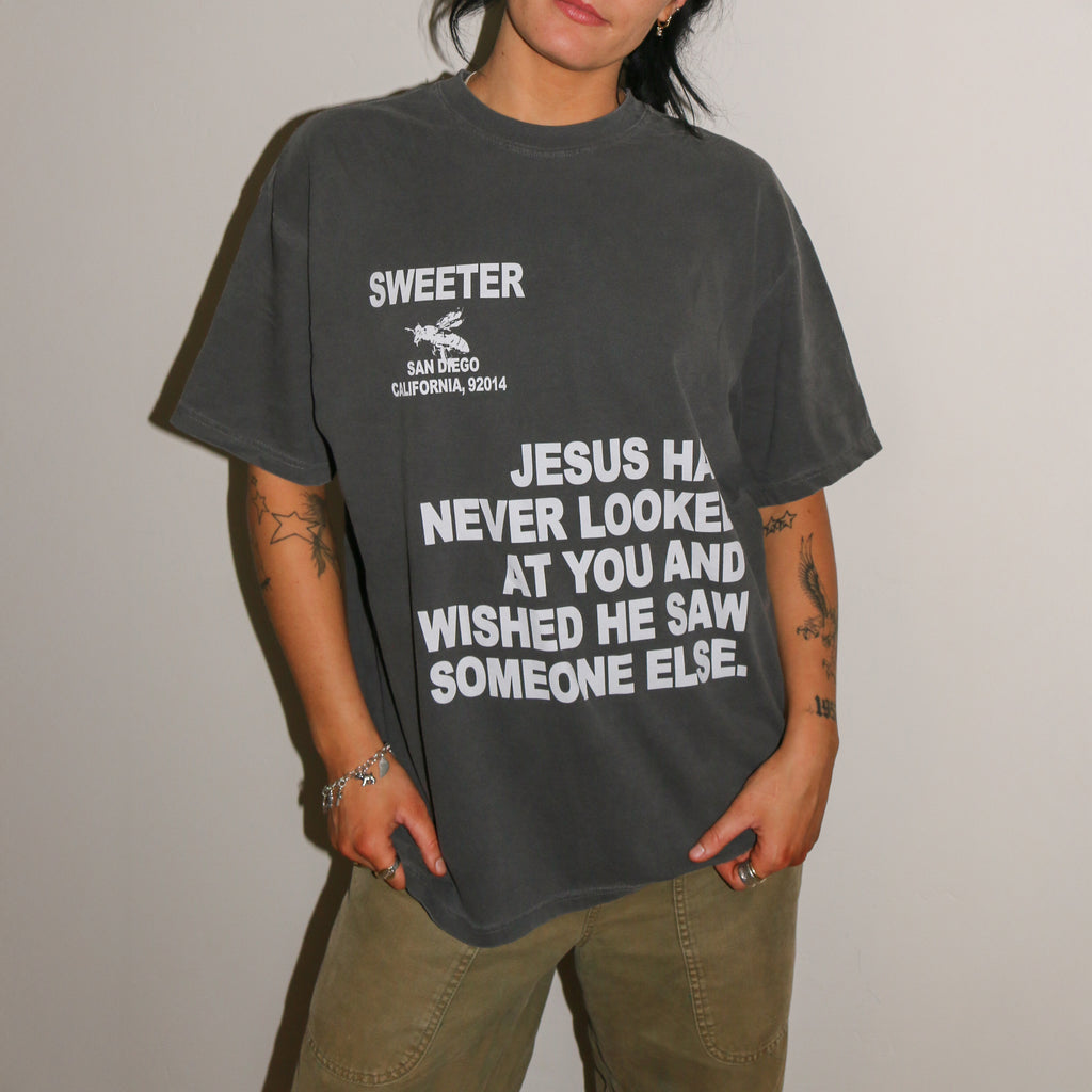 A person wearing a dark gray washed graphic t-shirt with white text that reads 'SWEETER SAN DIEGO CALIFORNIA, 2024' and 'JESUS HAS NEVER LOOKED AT YOU AND WISHED HE SAW SOMEONE ELSE.', paired with olive green cargo pants, standing against a plain light gray wall.