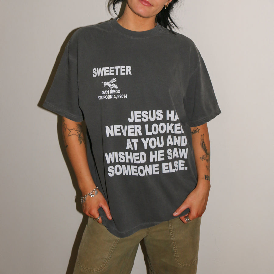 A person wearing a dark gray washed graphic t-shirt with white text that reads 'SWEETER SAN DIEGO CALIFORNIA, 2024' and 'JESUS HAS NEVER LOOKED AT YOU AND WISHED HE SAW SOMEONE ELSE.', paired with olive green cargo pants, standing against a plain light gray wall.