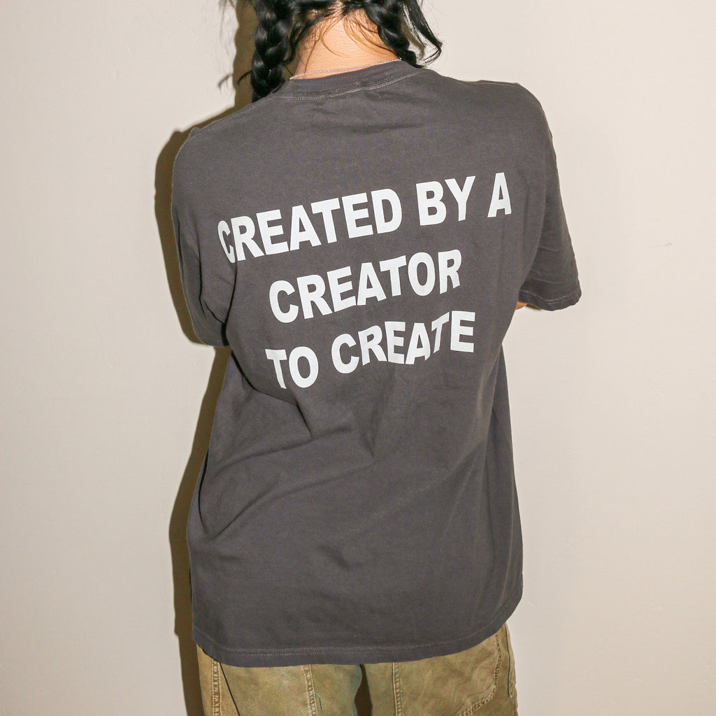 A person shown from behind wearing a dark gray t-shirt with the phrase 'CREATED BY A CREATOR TO CREATE' printed in bold white capital letters on the back, paired with olive green pants against a plain off-white wall.