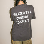 A person shown from behind wearing a dark gray t-shirt with the phrase 'CREATED BY A CREATOR TO CREATE' printed in bold white capital letters on the back, paired with olive green pants against a plain off-white wall.