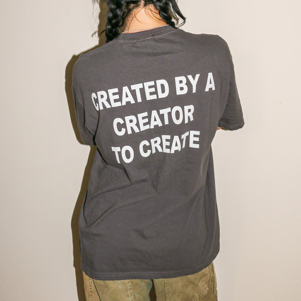 A person shown from behind wearing a dark gray t-shirt with the phrase 'CREATED BY A CREATOR TO CREATE' printed in bold white capital letters on the back, paired with olive green pants against a plain off-white wall.