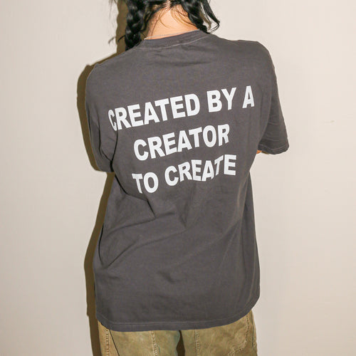A person shown from behind wearing a dark gray t-shirt with the phrase 'CREATED BY A CREATOR TO CREATE' printed in bold white capital letters on the back, paired with olive green pants against a plain off-white wall.