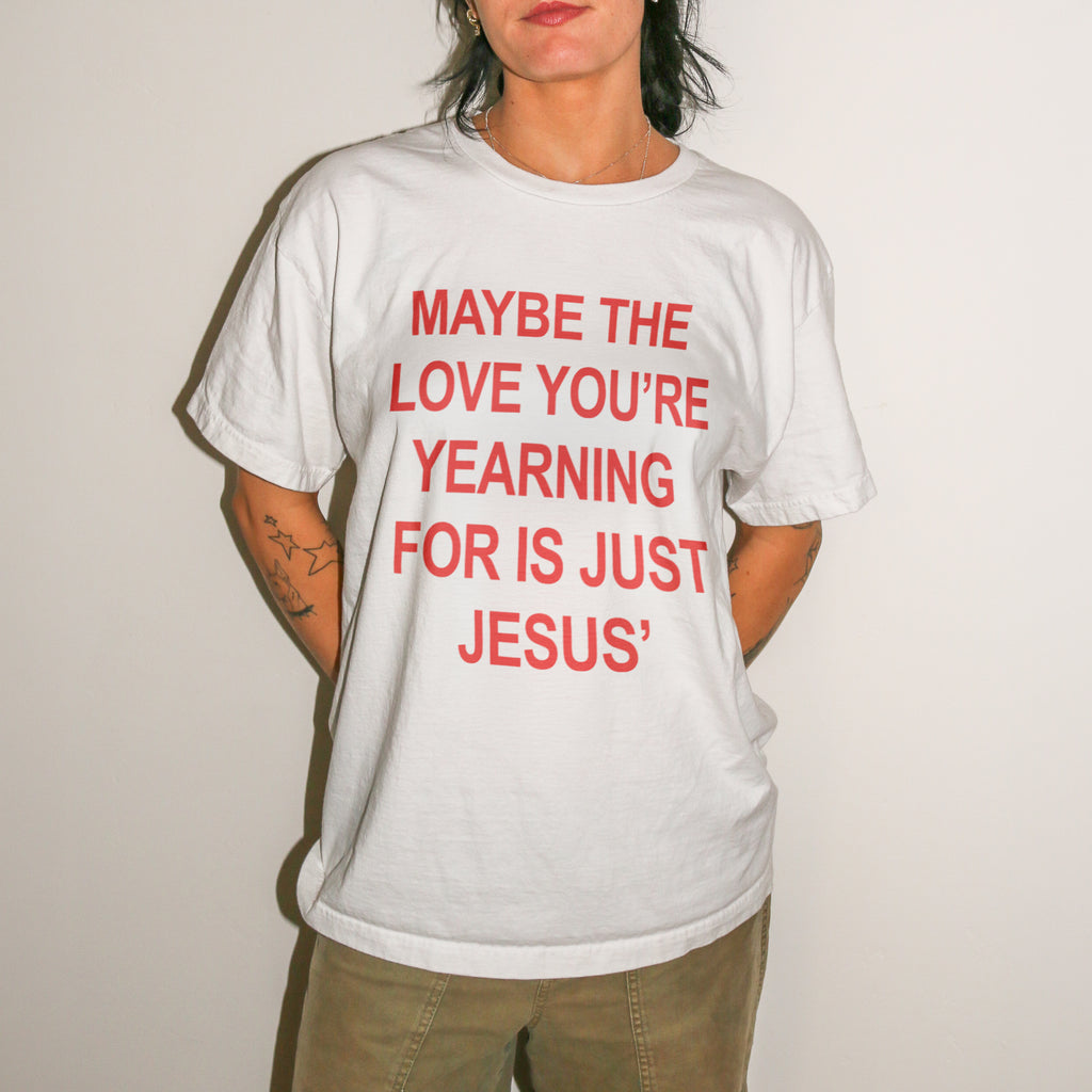 A person wearing a white short-sleeve graphic T-shirt with bold red text reading 'MAYBE THE LOVE YOU'RE YEARNING FOR IS JUST JESUS' and khaki pants, standing against a plain light wall.