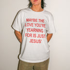 A person wearing a white short-sleeve graphic T-shirt with bold red text reading 'MAYBE THE LOVE YOU'RE YEARNING FOR IS JUST JESUS' and khaki pants, standing against a plain light wall.