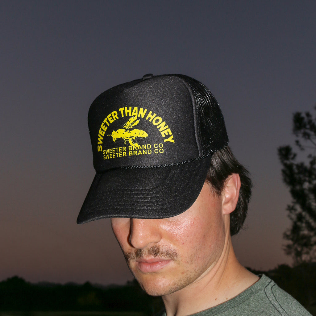 A man wearing a black trucker hat with yellow embroidered text reading 'SWEETER THAN HONEY' and 'SWEETER BRAND CO' on the front, photographed outdoors against a dusk sky.