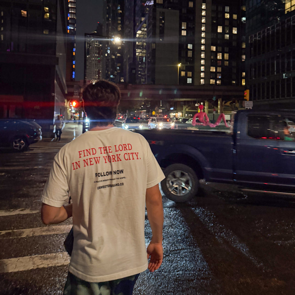 A man standing on a wet city street at night wearing a cream-colored t-shirt with red text on the back that reads 'FIND THE LORD IN NEW YORK CITY. FOLLOW NOW' and 'COMME des GARÇONS'.