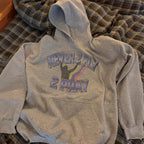 A heather gray pullover hoodie laid flat on a plaid bedspread. The front has a graphic with the words “NEVER 2 FLY 2 PRAY” in outlined lettering above and below a black silhouette of a person praying with a rainbow arc.