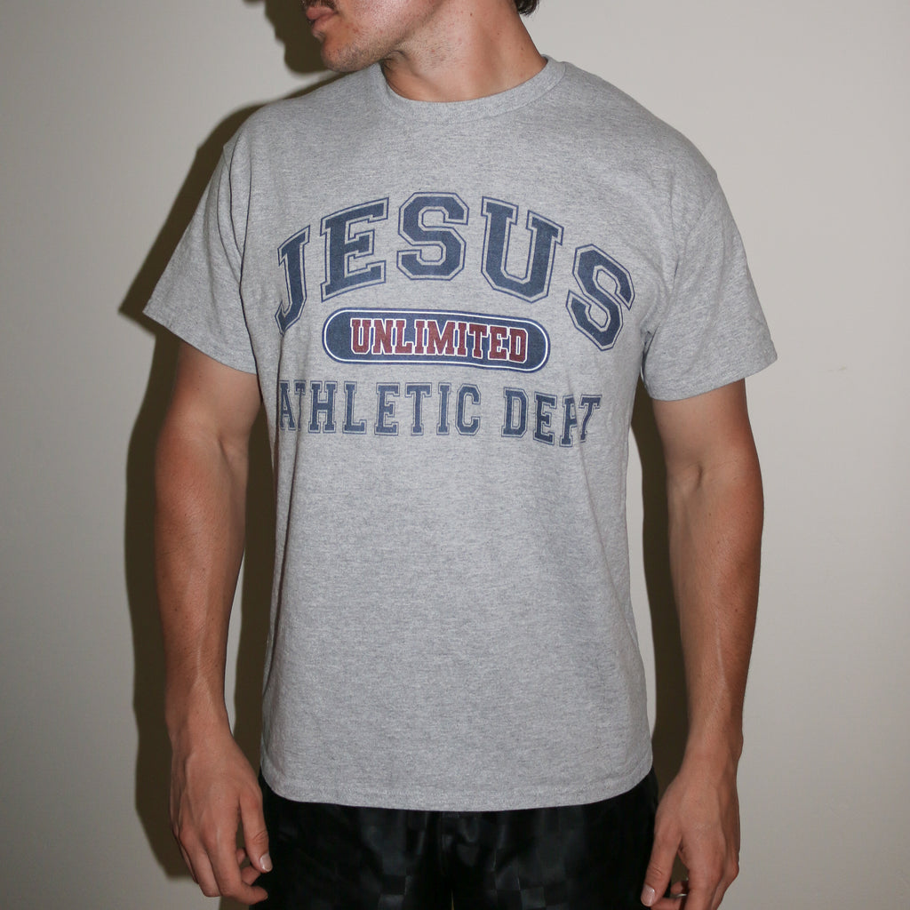 A man wearing a heather gray t-shirt with a collegiate-style graphic that reads 'JESUS UNLIMITED ATHLETIC DEPT' in navy blue and red, paired with black pants against a plain beige wall.