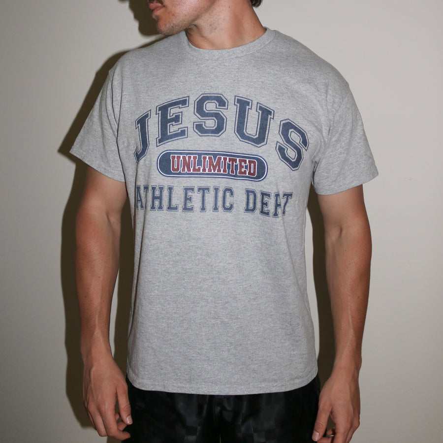 A man wearing a heather gray t-shirt with a collegiate-style graphic that reads 'JESUS UNLIMITED ATHLETIC DEPT' in navy blue and red, paired with black pants against a plain beige wall.