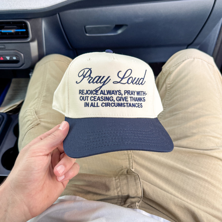 A person sitting in a car holds a beige and navy blue baseball cap with the text 'Pray Loud REJOICE ALWAYS, PRAY WITH-OUT CEASING, GIVE THANKS IN ALL CIRCUMSTANCES' embroidered on the front.
