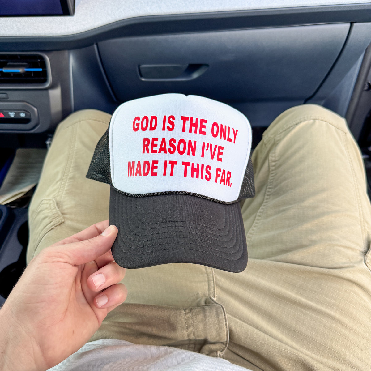 A person sitting in a car holds a trucker hat with a white front panel, black mesh sides, and red text that reads, 'GOD IS THE ONLY REASON I'VE MADE IT THIS FAR.'.