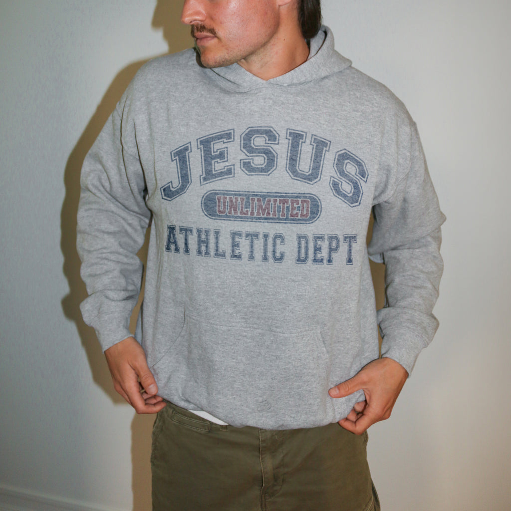 A man wearing a heather gray pullover hoodie with 'JESUS UNLIMITED ATHLETIC DEPT' printed in navy blue on the front, paired with olive green pants, standing against a plain light gray wall.