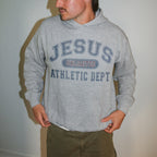 A man wearing a heather gray pullover hoodie with 'JESUS UNLIMITED ATHLETIC DEPT' printed in navy blue on the front, paired with olive green pants, standing against a plain light gray wall.