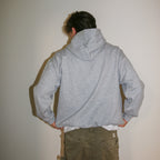 A person shown from behind wearing a light gray heathered pullover hoodie and olive green pants, standing against a plain off-white wall.