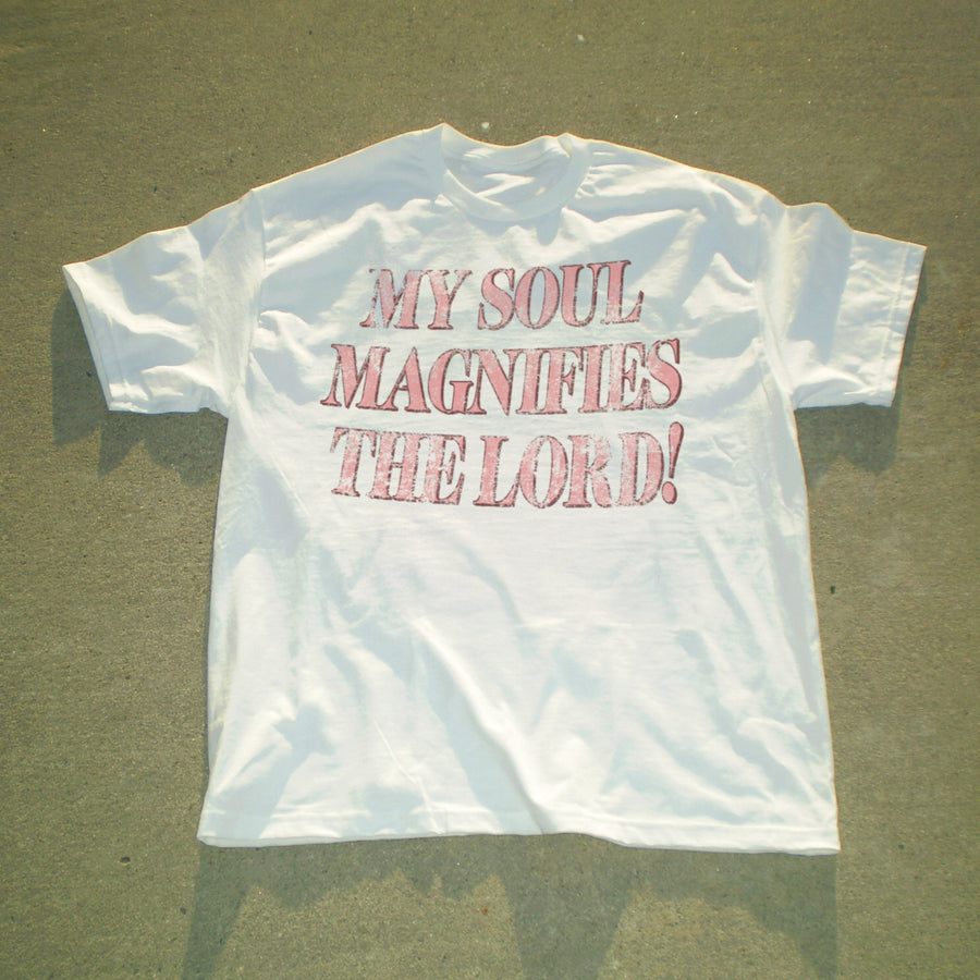 A white short-sleeve t-shirt laid flat on a textured gray surface, featuring the text 'MY SOUL MAGNIFIES THE LORD!' printed in a distressed pink font across the chest.