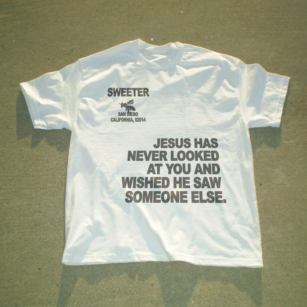 A white short-sleeve t-shirt laid flat on a gray concrete surface, featuring black printed text and a small bee graphic. The text reads 'SWEETER SAN DIEGO CALIFORNIA, 92914' above a larger quote: 'JESUS HAS NEVER LOOKED AT YOU AND WISHED HE SAW SOMEONE ELSE.