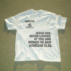 A white short-sleeve t-shirt laid flat on a gray concrete surface, featuring black printed text and a small bee graphic. The text reads 'SWEETER SAN DIEGO CALIFORNIA, 92914' above a larger quote: 'JESUS HAS NEVER LOOKED AT YOU AND WISHED HE SAW SOMEONE ELSE.