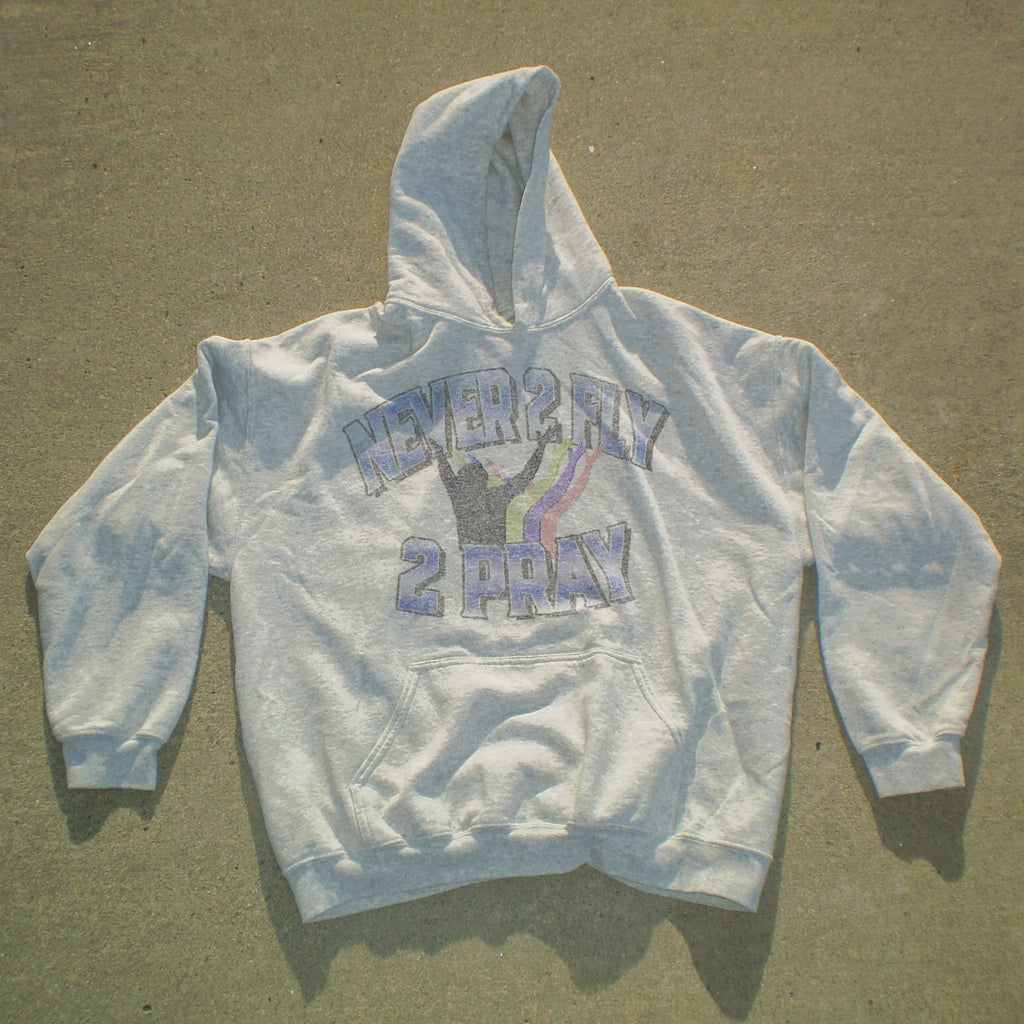 A light gray pullover hoodie laid flat on a concrete surface, featuring a distressed graphic on the chest that reads 'NEVER 2 HAY 2 PRAY' in purple and black with a purple ribbon design.