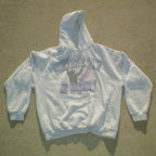 A light gray pullover hoodie laid flat on a concrete surface, featuring a distressed graphic on the chest that reads 'NEVER 2 HAY 2 PRAY' in purple and black with a purple ribbon design.